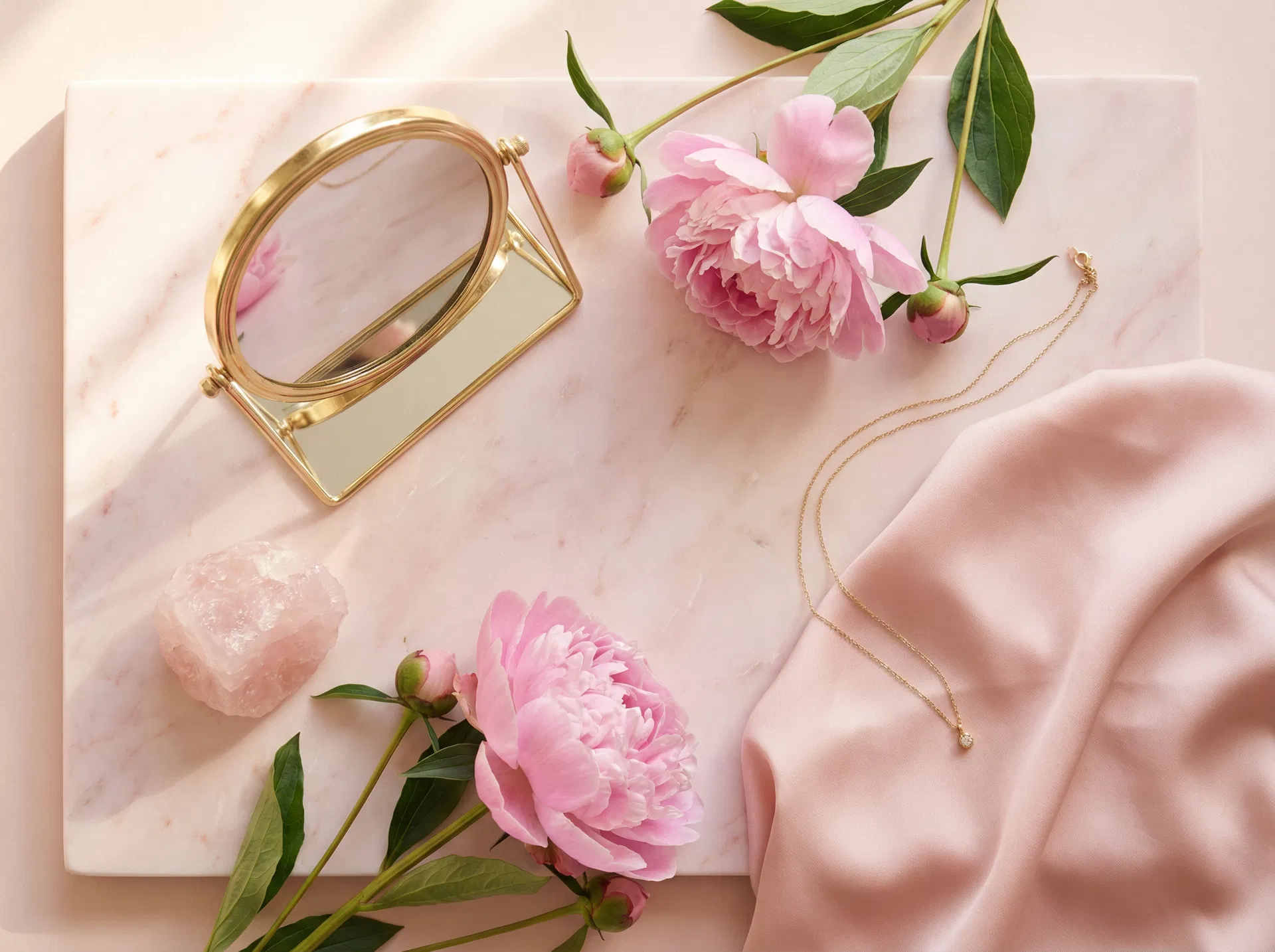 Elegant flat-lay of pink peonies, gold mirror, rose quartz crystal and silk fabric representing Glow Goddess LLC beauty and wellness services