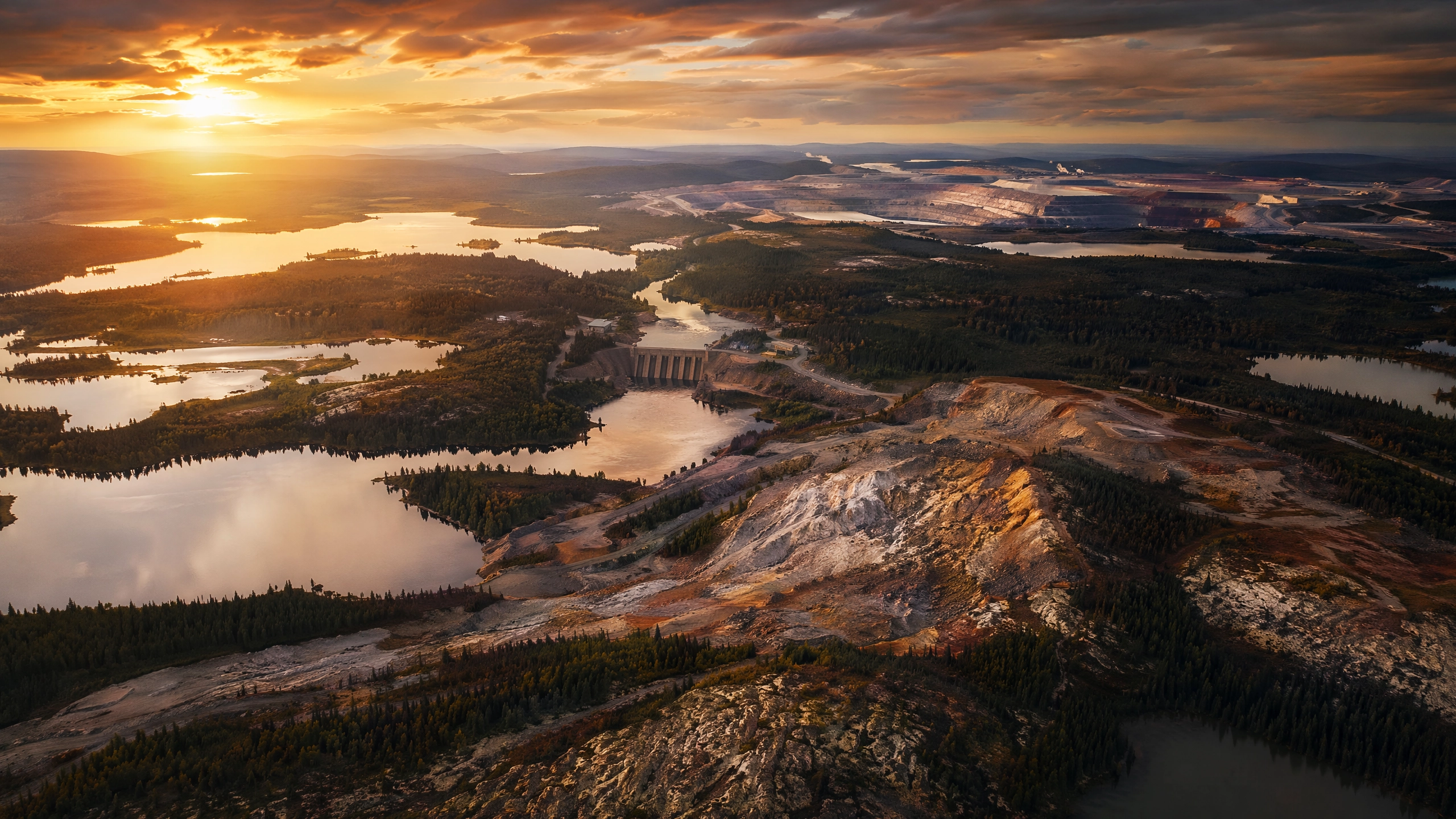 Aerial view of the Canadian Shield at golden hour showing boreal forest, pristine lakes, a hydroelectric dam, and mining operations