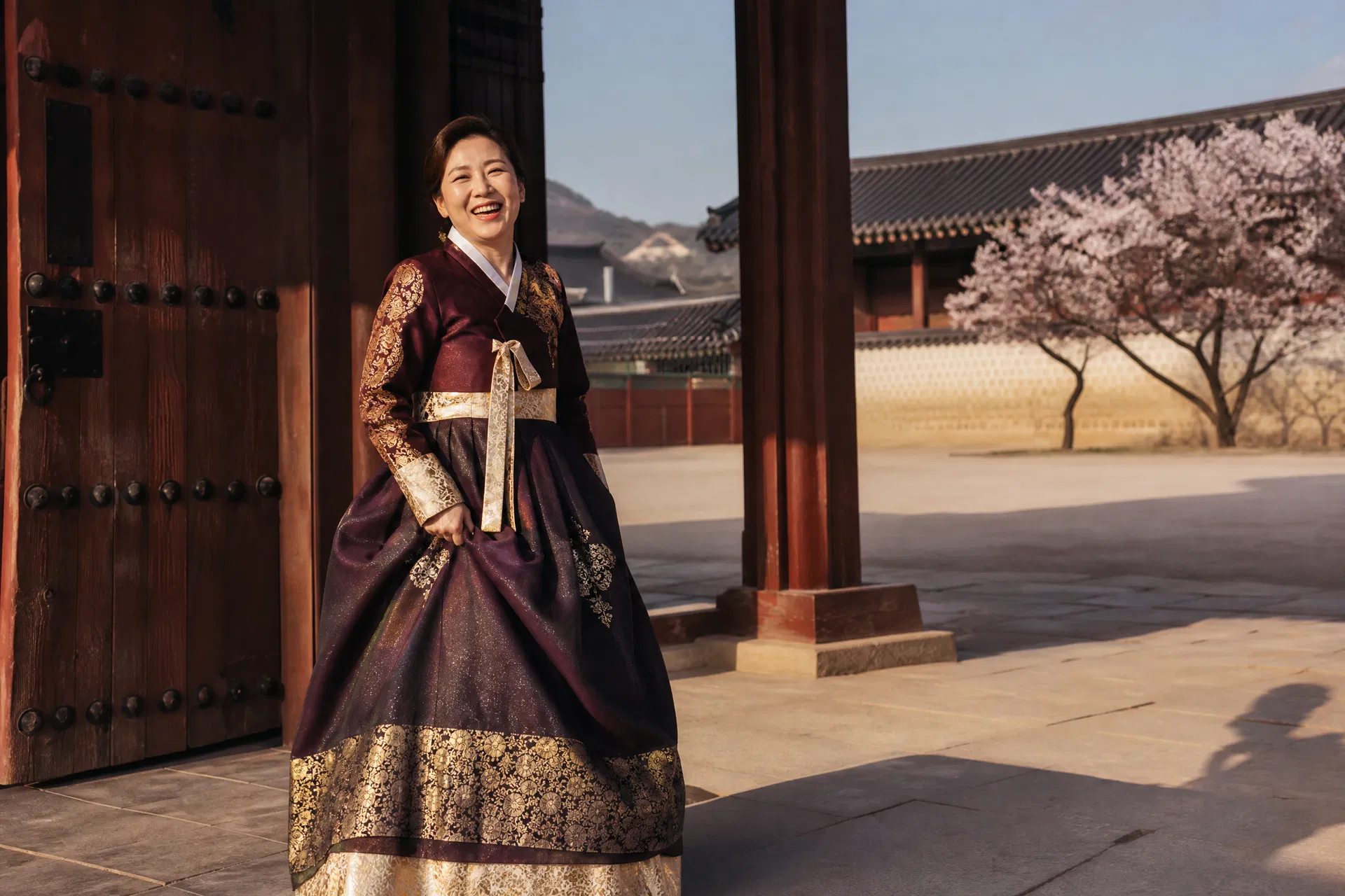 A woman in her late 40s wearing a beautifully fitted traditional Korean hanbok in deep burgundy and gold, laughing naturally in front of the ornate wooden gates of Gyeongbokgung Palace, cherry blossoms visible in the background.