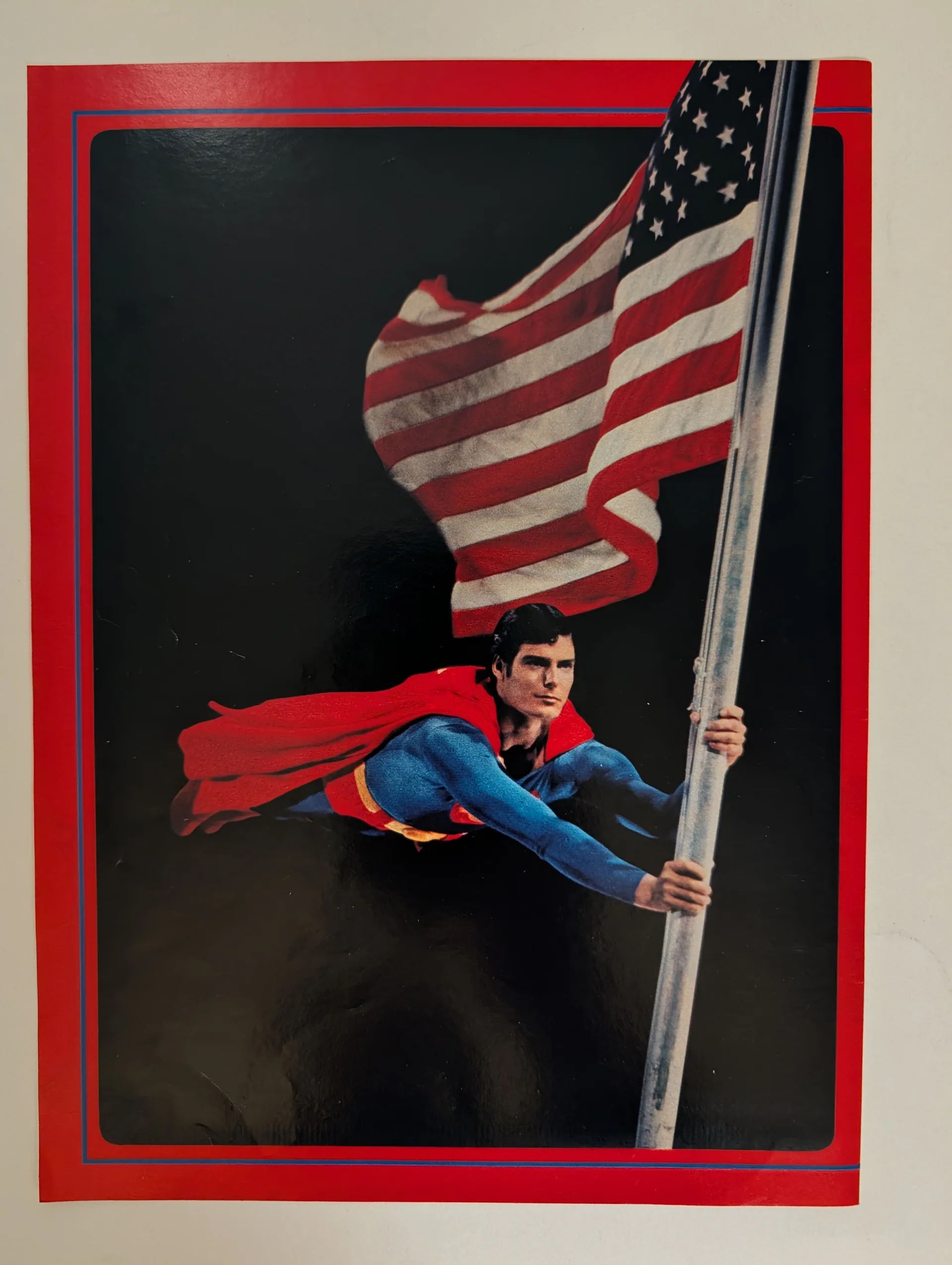 Superman flag poster: the figure in flight gripping a flagpole, U.S. flag billowing above, black background, red border.
