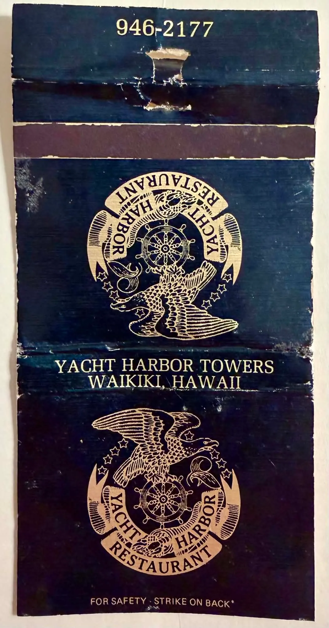 A flattened, vintage matchbook cover from the Yacht Harbor Restaurant. The exterior features a dark nautical blue background with a metallic gold logo of an eagle perched on a ship's wheel, flanked by stars and banners reading "YACHT HARBOR RESTAURANT". Gold text reads "YACHT HARBOR TOWERS / WAIKIKI, HAWAII", "946-2177", and "FOR SAFETY - STRIKE ON BACK*". The interior is printed on white cardstock with a wavy dark blue border, advertising "Gourmet Seafood & Continental Cuisine" and "Entertainment from 8 p.m." The interior is heavily stained with a large, organic brown liquid spill. The manufacturer's imprint reads "Adv. Spec. Hawaii, Honolulu, Hi-SM".