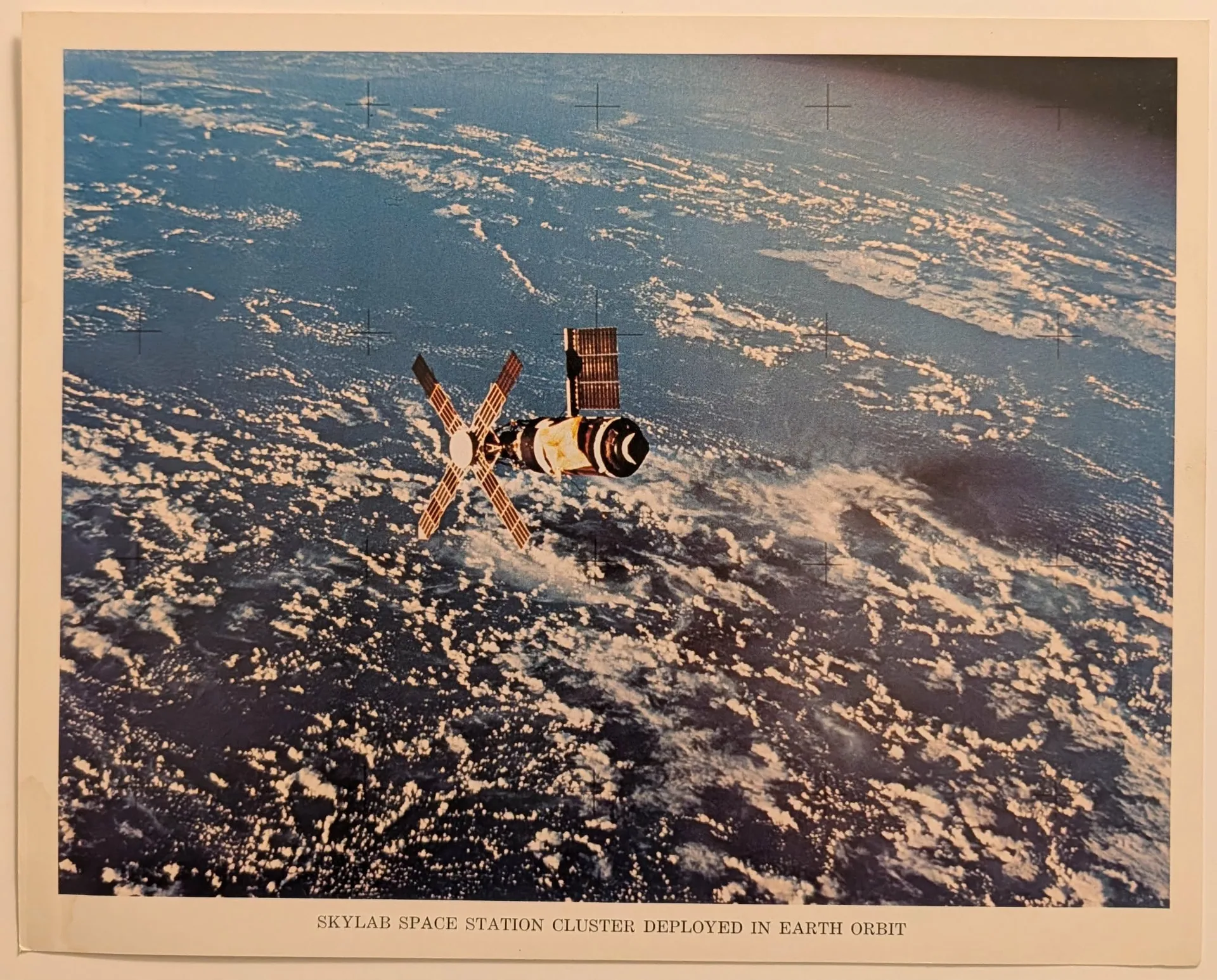 A color photograph of the Skylab space station orbiting Earth, set against a backdrop of dense white clouds and deep blue ocean. The station appears visibly damaged, missing one of its large main solar panels on its side. A makeshift, gold-colored parasol sunshade is deployed over the main cylindrical body of the station. At the front, four smaller solar panels extend in a cross or windmill formation. The image contains faint, evenly spaced black crosshairs (reseau marks) typical of scientific space cameras. Below the photograph, a printed caption in a serif font reads: "SKYLAB SPACE STATION CLUSTER DEPLOYED IN EARTH ORBIT".