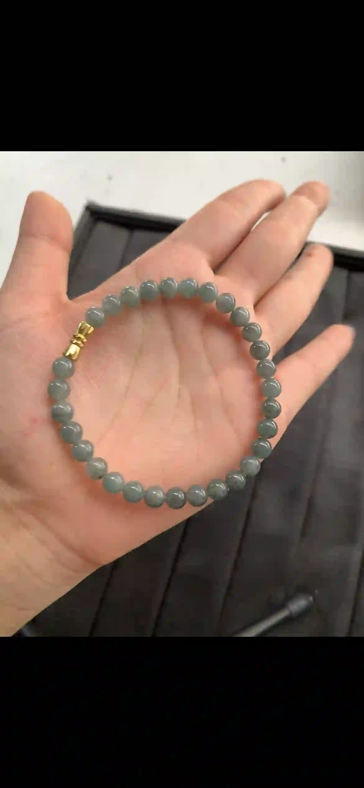 A sleek single-strand bracelet of evenly matched round jadeite beads, featuring a cool translucent greyish-green base with natural wispy dark inclusions, finished with a polished gold-toned clasp. | Myanmar Jadeite reference #11190001