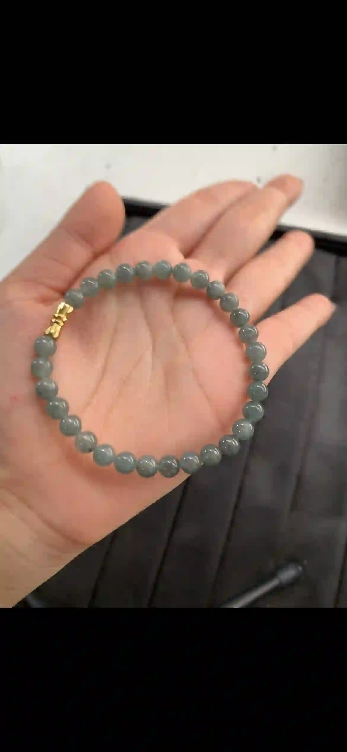 A sleek single-strand bracelet of evenly matched round jadeite beads, featuring a cool translucent greyish-green base with natural wispy dark inclusions, finished with a polished gold-toned clasp., view 2 | Myanmar Jadeite reference #11190001