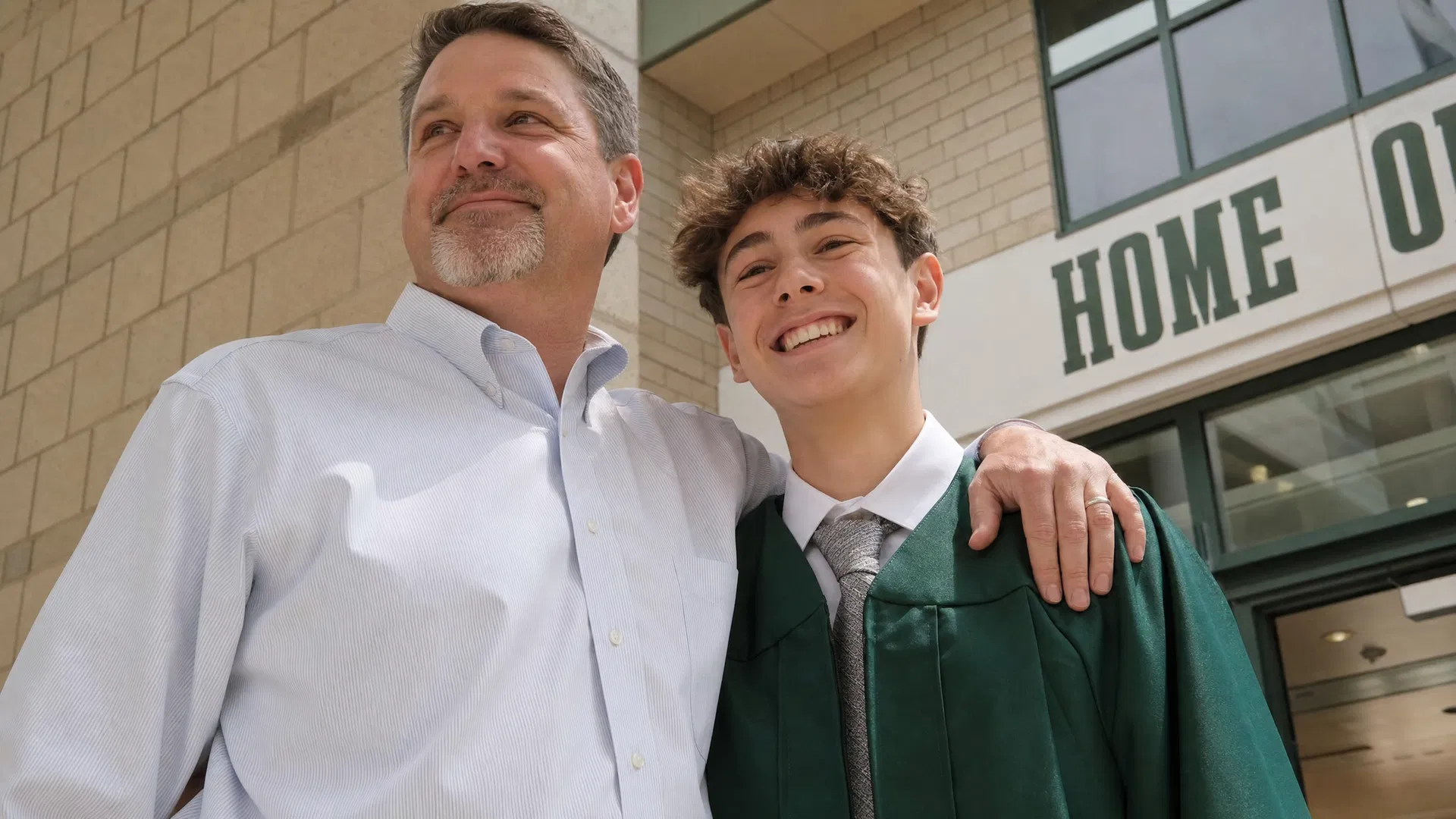 Dad with his arm around his son in graduation gown outside the school, both smiling