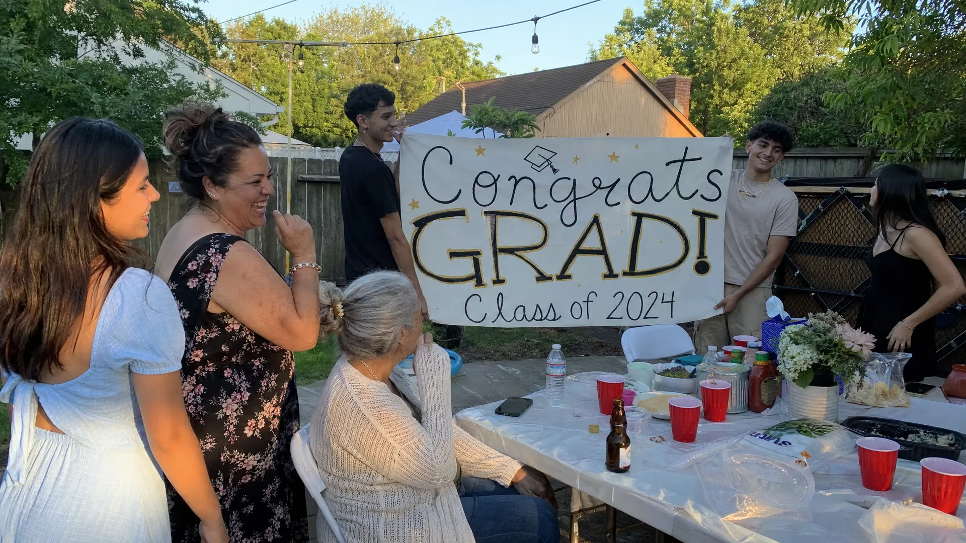 Backyard graduation party with family laughing around a table with a Congrats Grad banner