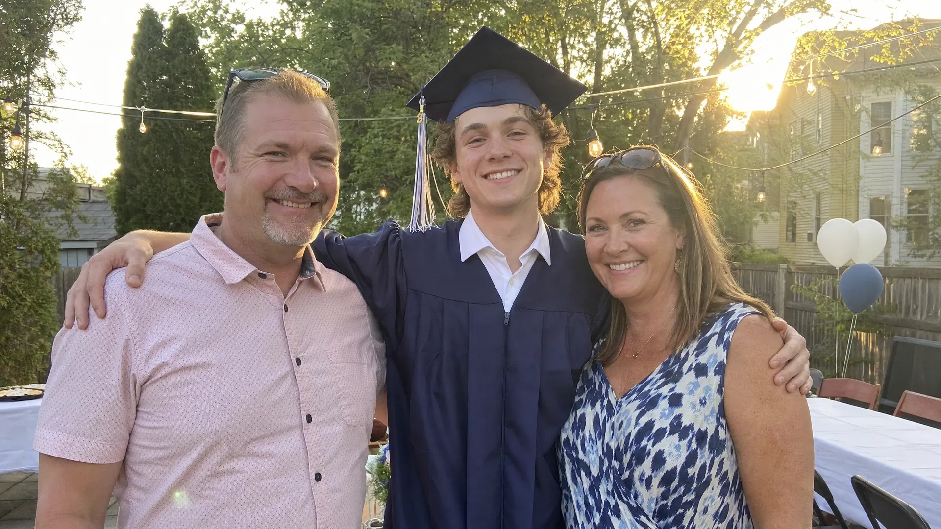 Both parents standing with their son in graduation cap and gown at a backyard graduation party, golden hour light