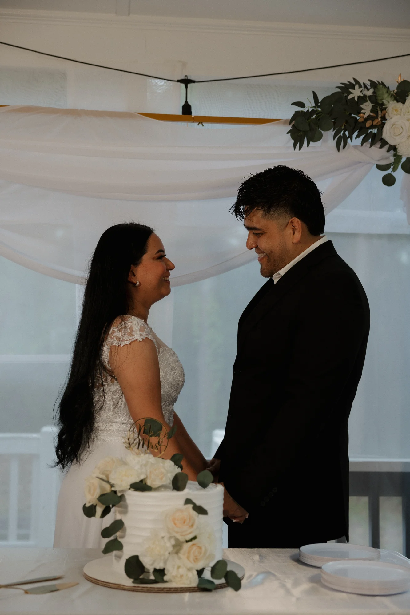 Smiling bride and groom holding hands at their intimate wedding reception next to a white wedding cake with floral decor.