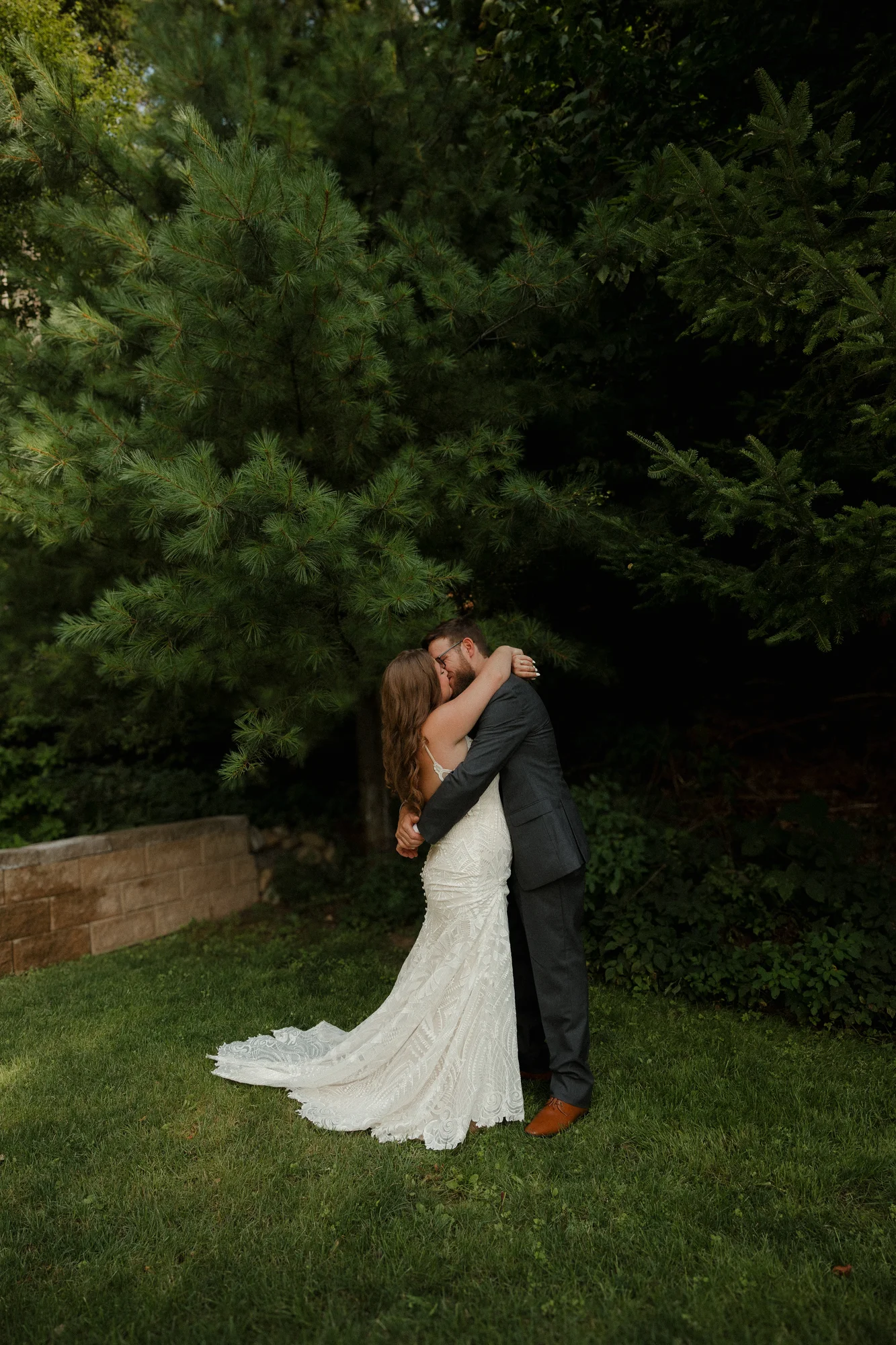Bride in lace gown and groom in suit share a romantic kiss outdoors, surrounded by deep green pine trees.
