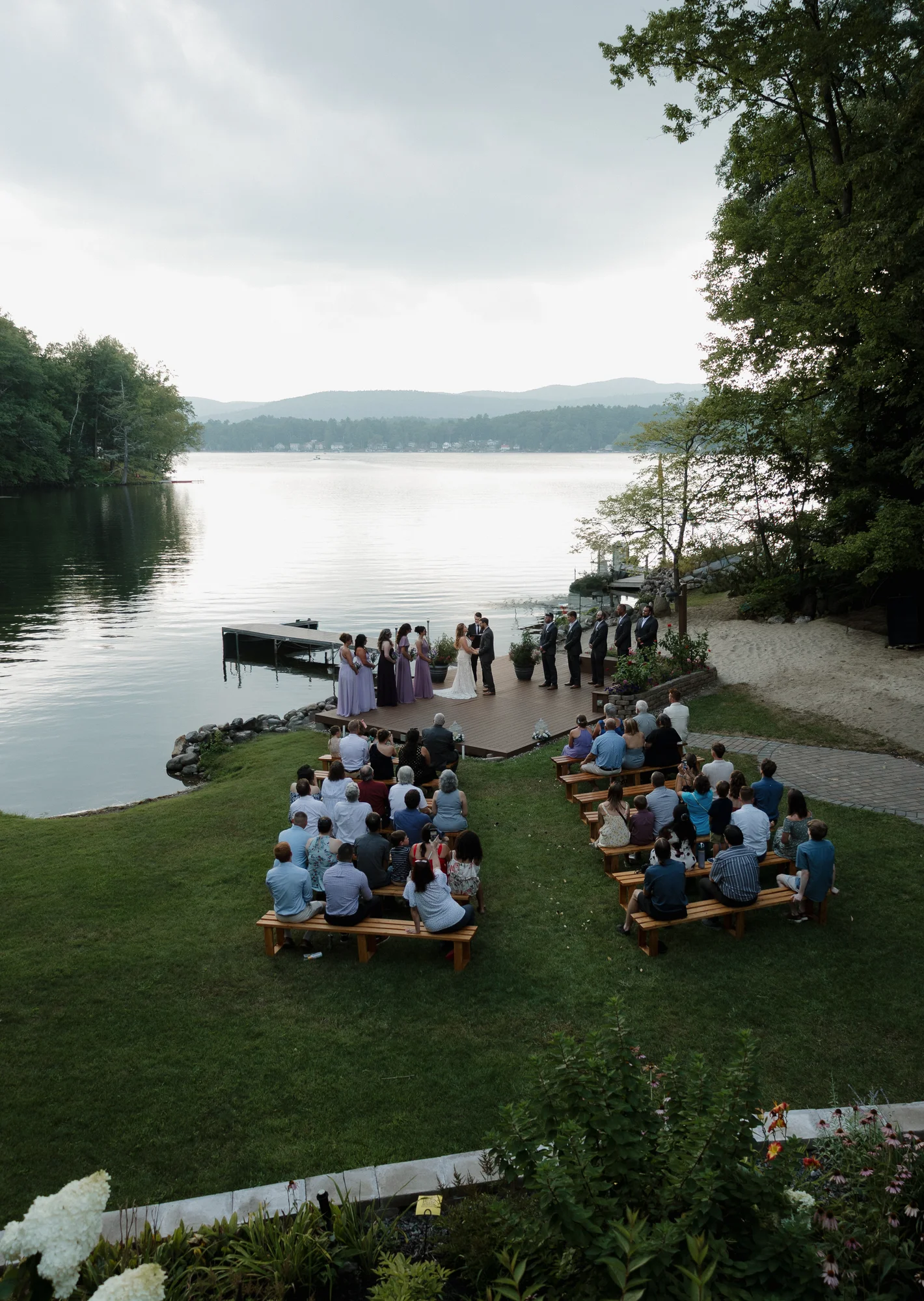 Overhead view of a wedding ceremony on a lake dock, with a couple, wedding party, and guests. Mountains in background.