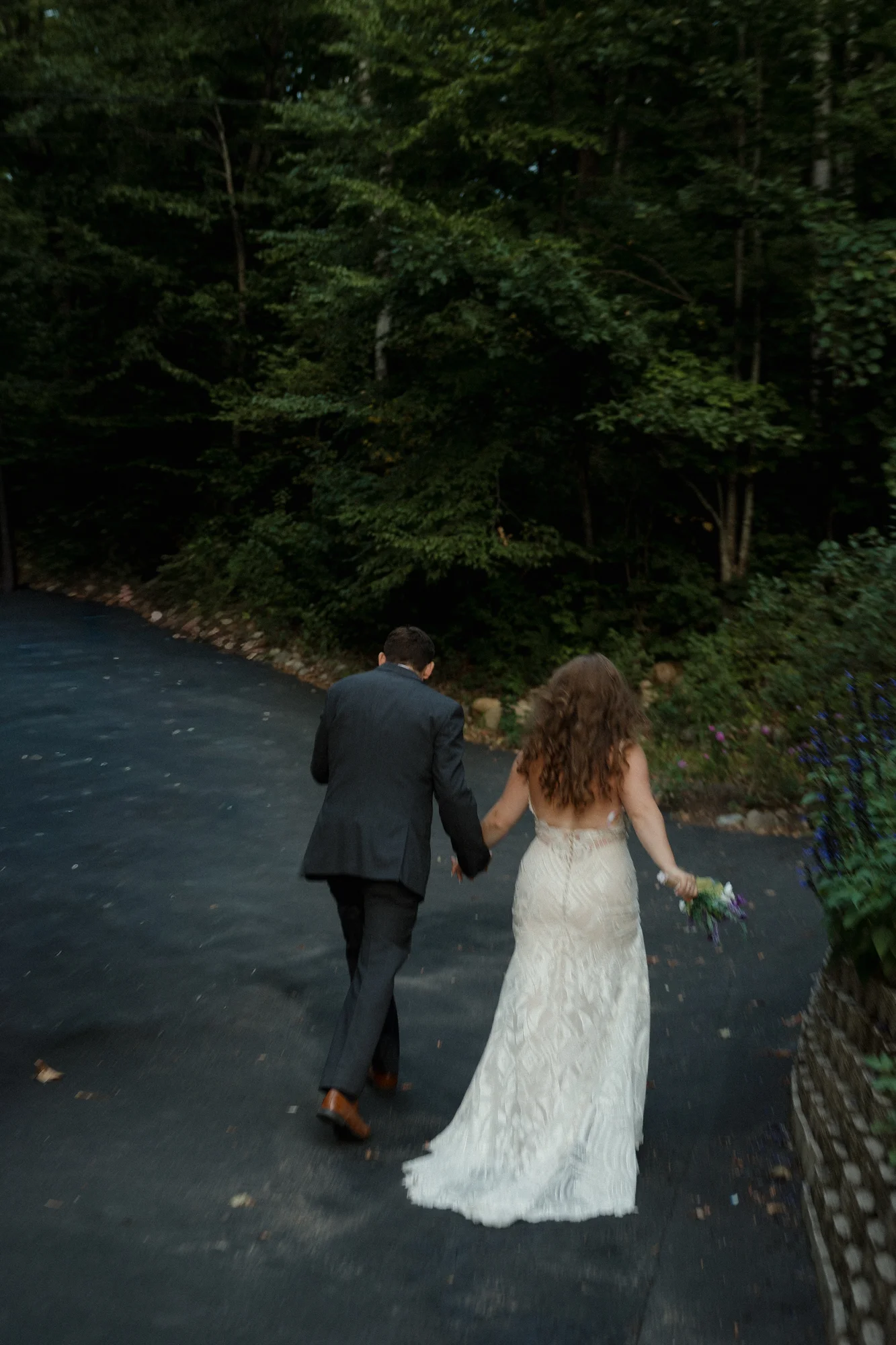 Newlywed couple, bride in a backless lace dress and groom in a suit, walking hand-in-hand on a path surrounded by trees.