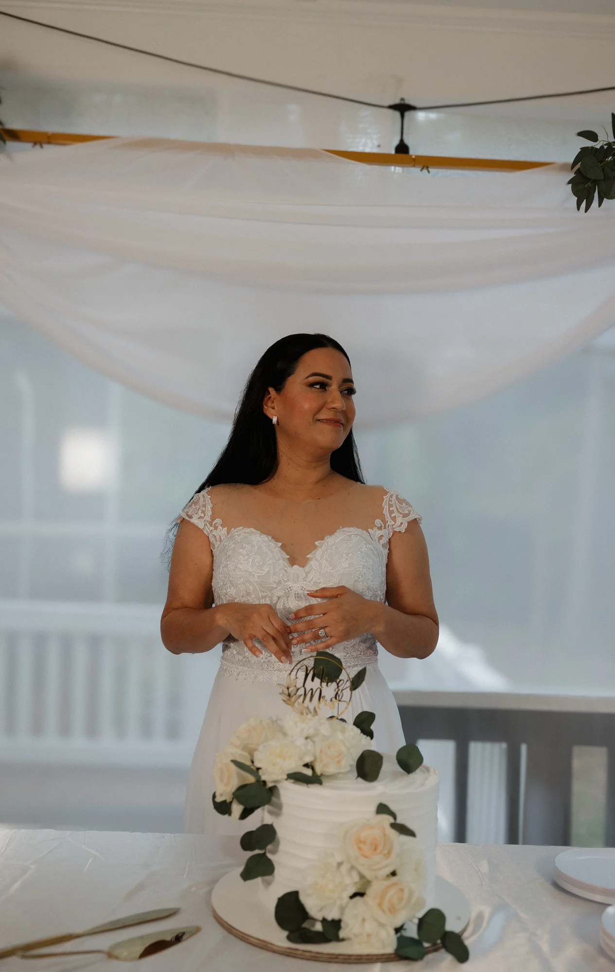A smiling bride in a white lace wedding dress stands next to a white wedding cake with floral decorations and a "Mr & Mrs" topper.