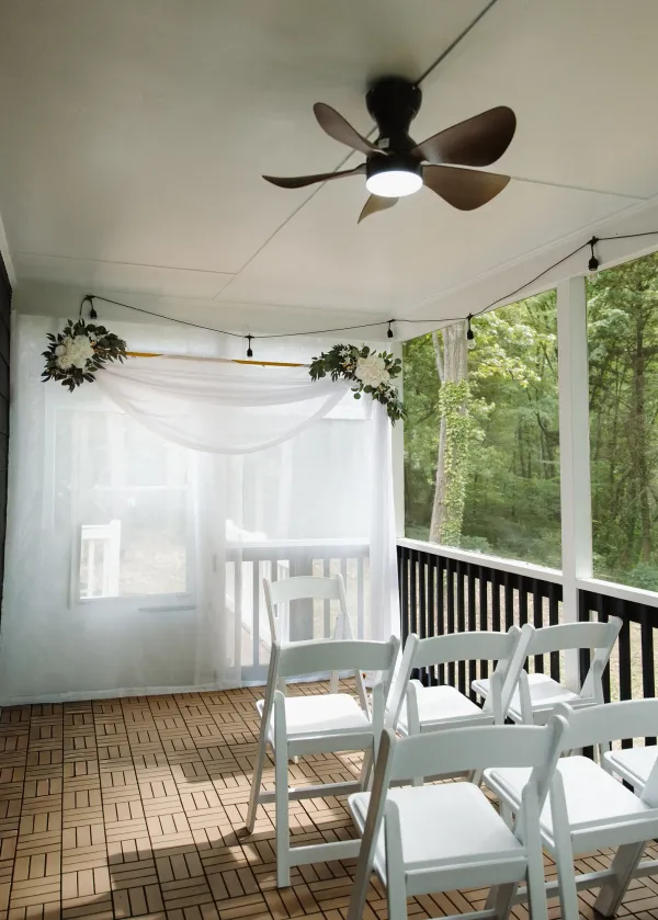 Outdoor wedding ceremony setup on a porch with white chairs, sheer white drapes, and floral decor, overlooking a forest.