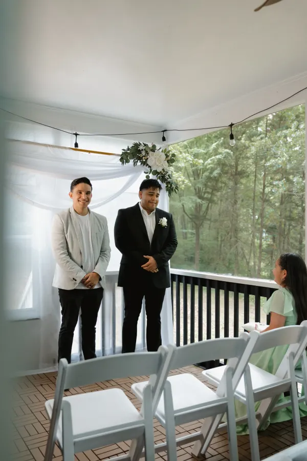 Two men, one in a light blazer and one in a black suit with a boutonniere, stand on a decorated balcony for a wedding.