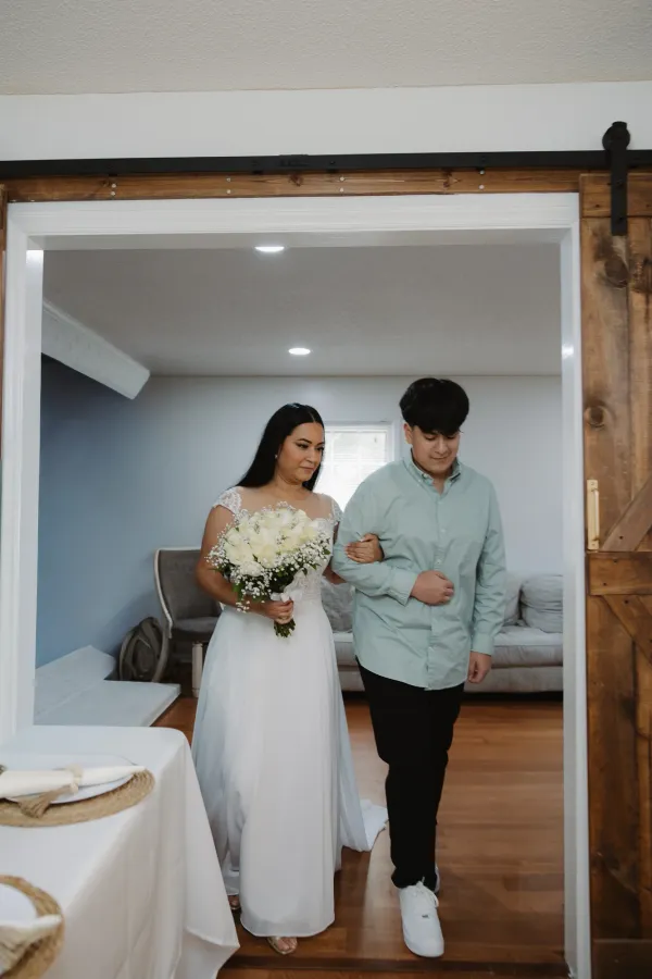 Bride in white dress holding white rose bouquet, escorted by a young man through a wooden doorway.