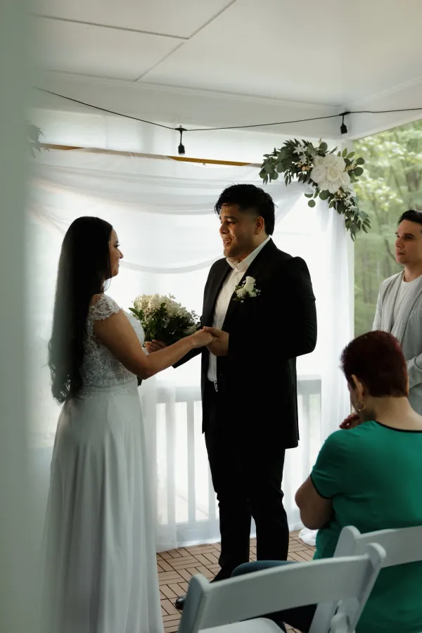 Bride and groom holding hands during their wedding ceremony, smiling at each other under a decorated arch.