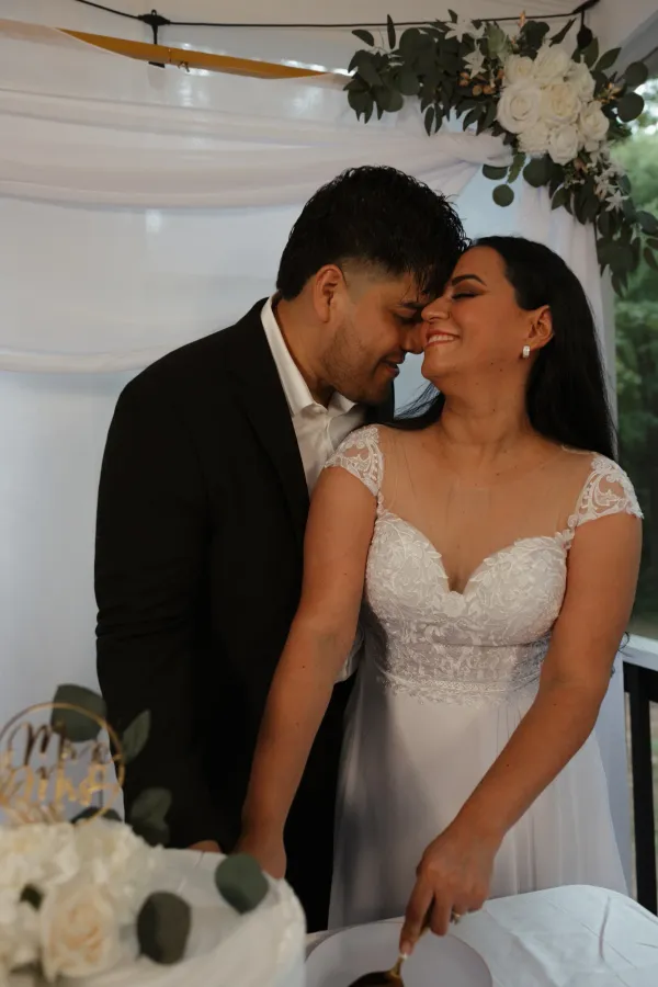 Bride and groom smiling, foreheads touching, cutting a white wedding cake with floral decoration.