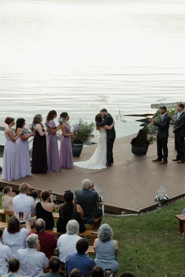 Newlyweds kissing on a wooden deck by a calm lake during their wedding ceremony, with guests watching.