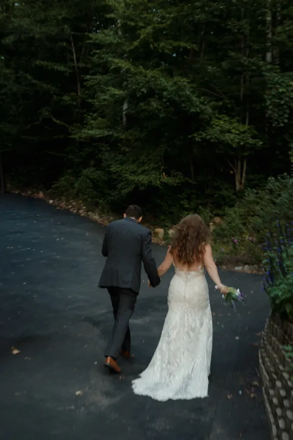 Newlywed couple, bride in a backless lace dress and groom in a suit, walking hand-in-hand on a path surrounded by trees.