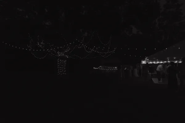 Outdoor wedding reception at night with string lights, a lit tent, and silhouettes of guests dancing under trees.