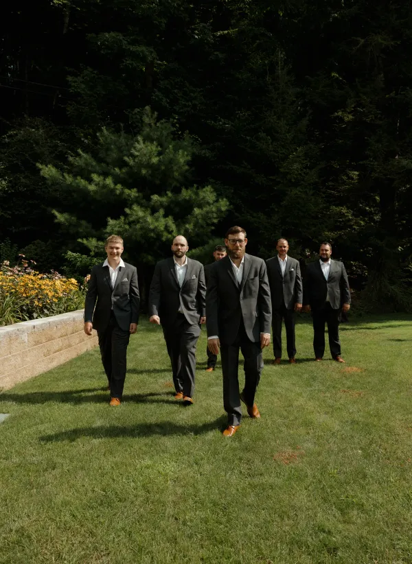 Five groomsmen in dark suits and brown shoes walk on a sunny green lawn with trees in the background.
