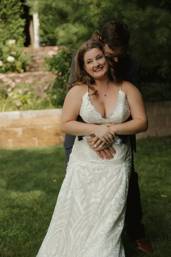 Bride in white lace dress smiling, embraced by groom from behind in a garden setting, showcasing their love and happiness.