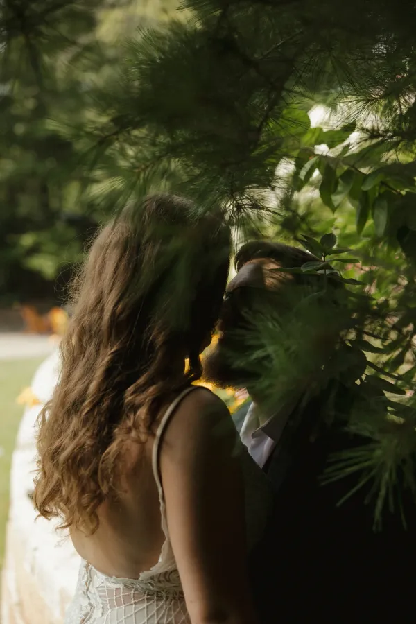Bride with long curly hair and groom with beard sharing an intimate moment, framed by green pine needles.