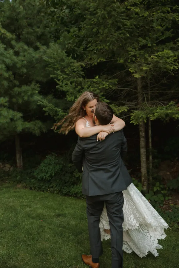 Groom lifting bride in a joyful embrace outdoors, surrounded by green trees, her long hair flowing.