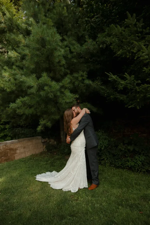 Bride in lace gown and groom in suit share a romantic kiss outdoors, surrounded by deep green pine trees.