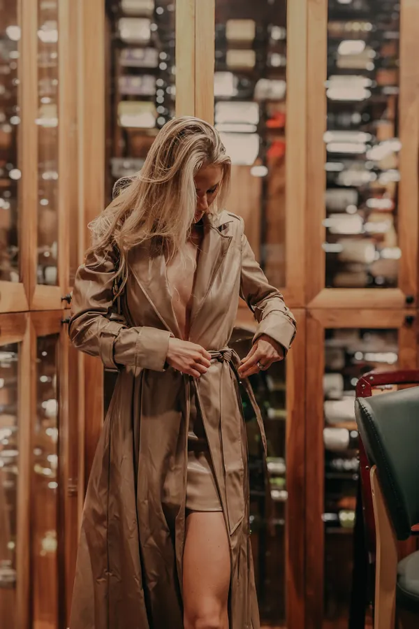 Blonde woman in a tan trench coat tying her belt in a dimly lit room with wine racks in the background, showcasing a natural and elegant lifestyle moment.