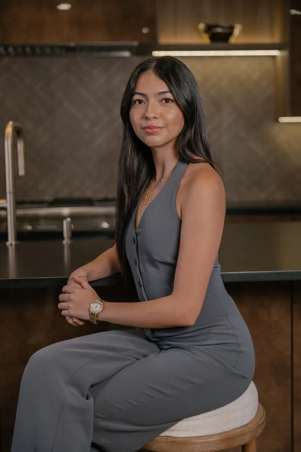 Professional headshot of a confident young woman, a Charlotte NC real estate agent, sitting at a modern kitchen counter during a branding photoshoot. She wears a stylish grey jumpsuit and a gold watch.