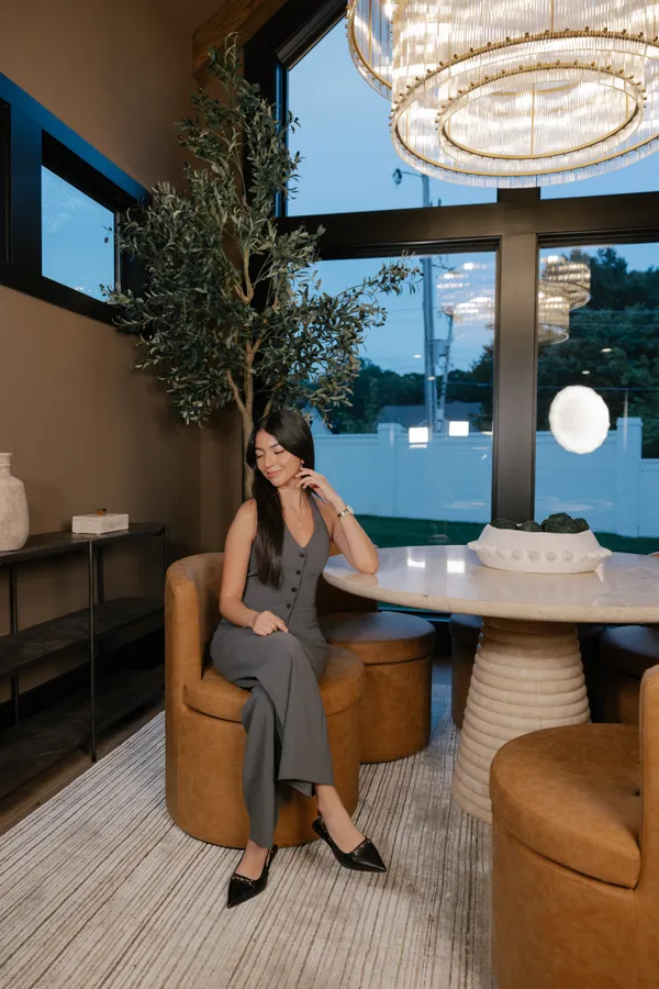 Professional headshot of a smiling female real estate agent in Charlotte NC, wearing a grey jumpsuit and sitting in a modern office with large windows and stylish decor.