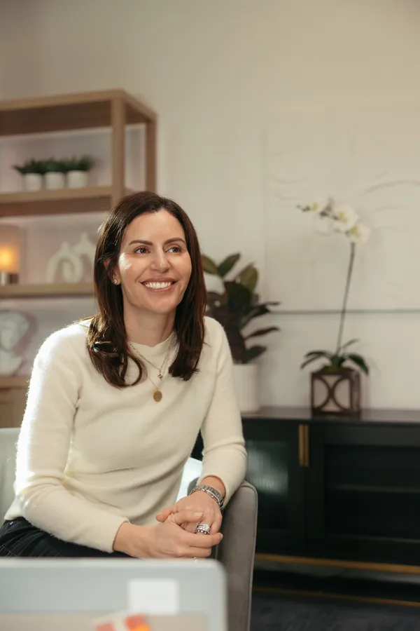 Smiling female podcast host in a cream sweater, looking engaged during a professional branding photoshoot in Charlotte NC. She is seated in a modern studio setting with a stylish bookshelf and decor in the background.