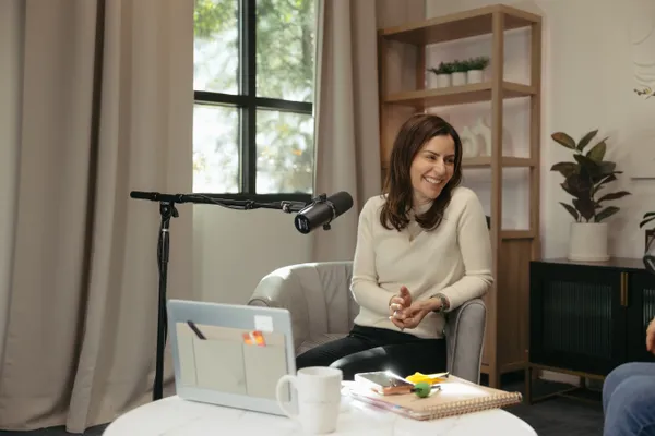 Smiling woman in a cream sweater sitting in a modern podcast studio with a microphone, laptop, and notes, captured during a branding photoshoot in Charlotte, NC.