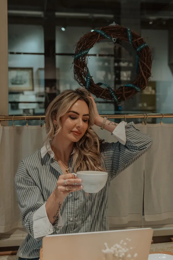 Blonde woman in striped shirt holding a white coffee cup, looking at a laptop in a cozy Charlotte NC coffee shop setting, with a decorative wreath in the background.
