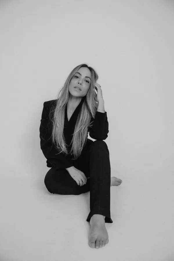 Black and white editorial portrait of a woman with long hair in a black suit, seated on the floor, looking directly at the camera. Her hand is gently touching her hair.