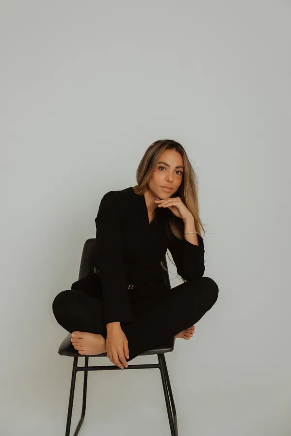 Editorial portrait of a woman in a black pantsuit sitting cross-legged on a chair, looking confidently at the camera, against a clean white background.