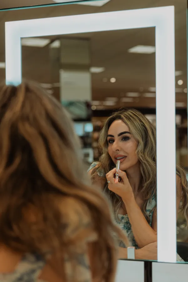 Blonde woman applying pink lipstick in a lighted vanity mirror, with her reflection visible.