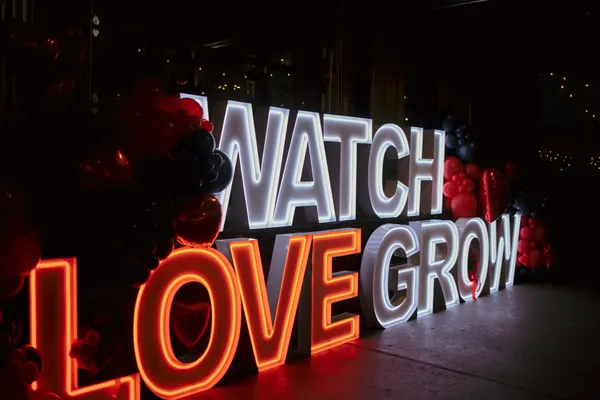 Illuminated marquee letters spelling "WATCH LOVE GROW" with clusters of red and black balloons around them, set against a dark background at a Galentine's event.