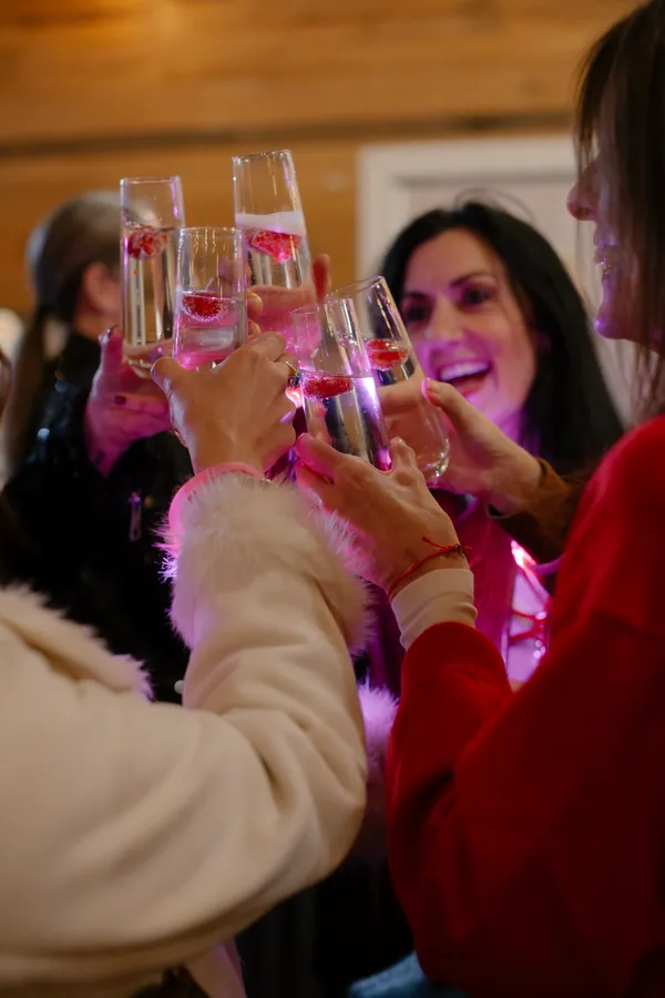 Close-up of several women's hands holding champagne flutes with raspberries, making a toast during a Galentine's Day celebration, with one woman smiling brightly in the background.