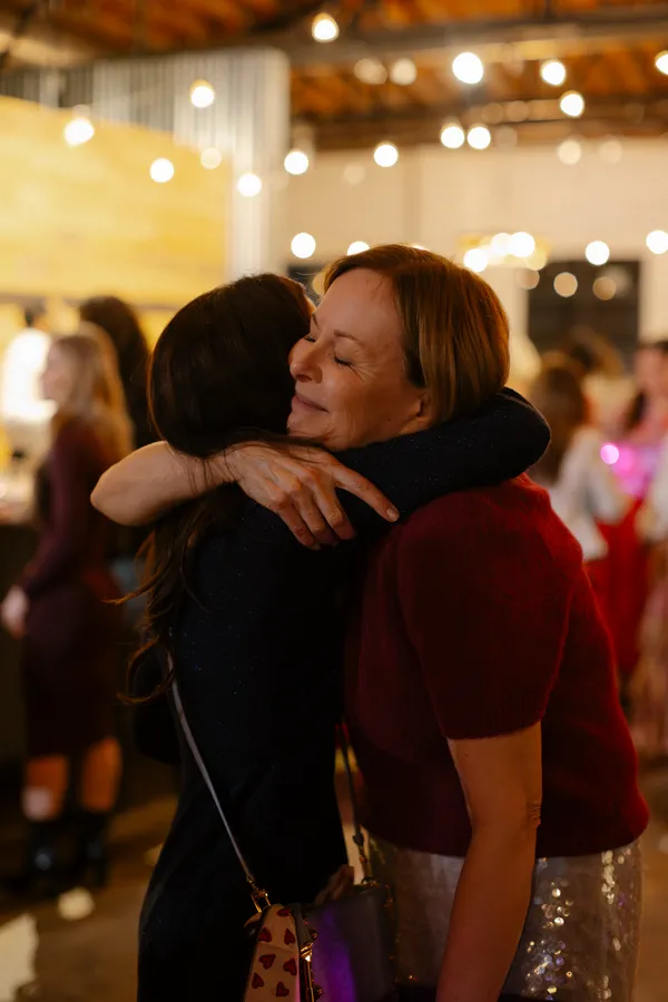 Two women embracing warmly at a Galentine's Day event, with one woman smiling with closed eyes, surrounded by soft bokeh lights, symbolizing friendship and connection.