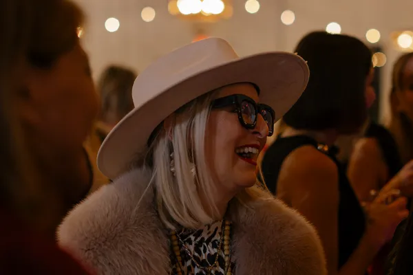 Close-up of a smiling woman with blonde hair, wearing a light-colored fedora hat, black glasses, red lipstick, and a fur coat, enjoying a Galentine's event in Charlotte.