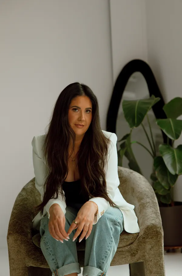 Professional woman with long dark hair, wearing a white blazer and jeans, sitting in an armchair for a personal branding photoshoot in Charlotte NC.