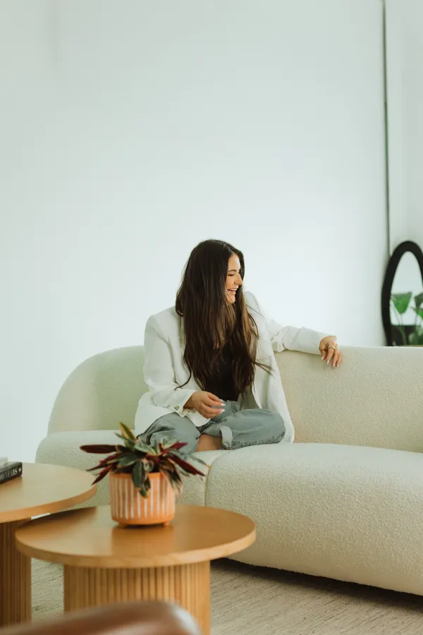 Smiling woman with long dark hair, wearing a white blazer and jeans, sitting comfortably on a light-colored sofa in a bright studio, perfect for personal branding photoshoots in Charlotte, NC.