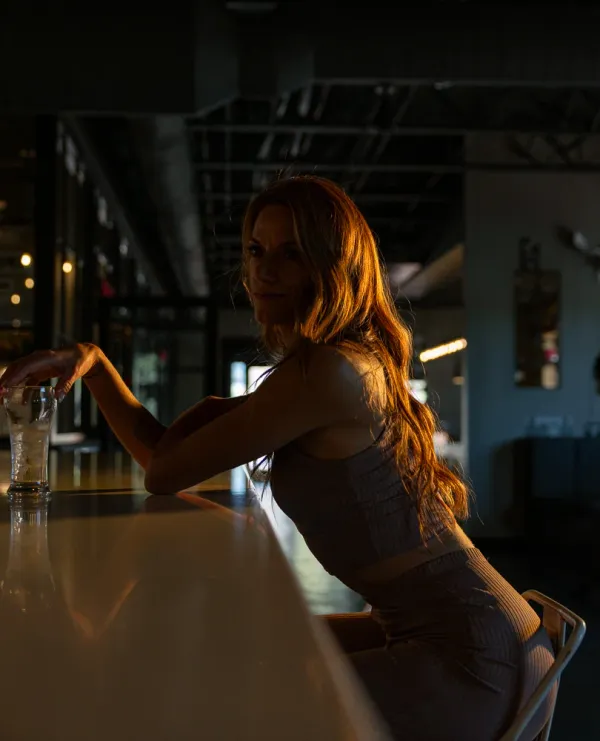 Woman with long, reddish-brown hair illuminated by golden hour light, leaning on a bar with a glass in hand, looking back over her shoulder with a subtle smile. The background is dark, showing industrial-style bar elements.