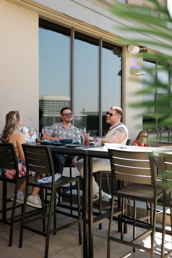 Guests laughing and enjoying cocktails at a summer rooftop event on a Charlotte restaurant patio, with city views in the background.