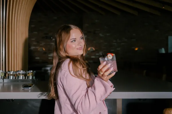 Young woman with long brown hair, wearing a pink jacket, holding a pink cocktail with lychee and strawberry garnish, looking up and smiling in a modern bar setting, ideal for lifestyle branding.
