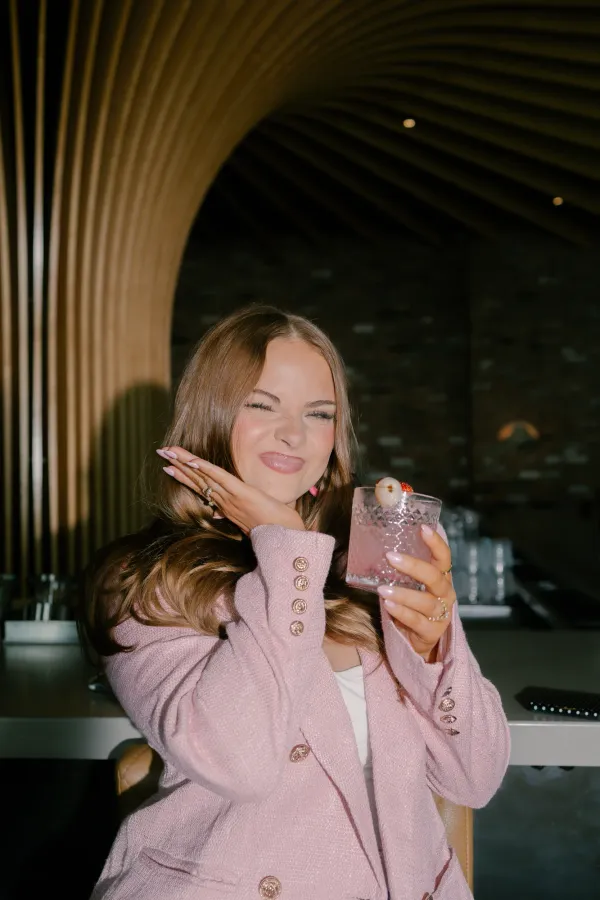 Young woman in pink jacket winking, holding a cocktail with lychee, playful expression.