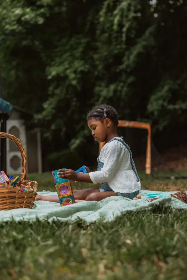 Young girl with braids playing with colorful magnetic tiles on a green blanket outdoors.