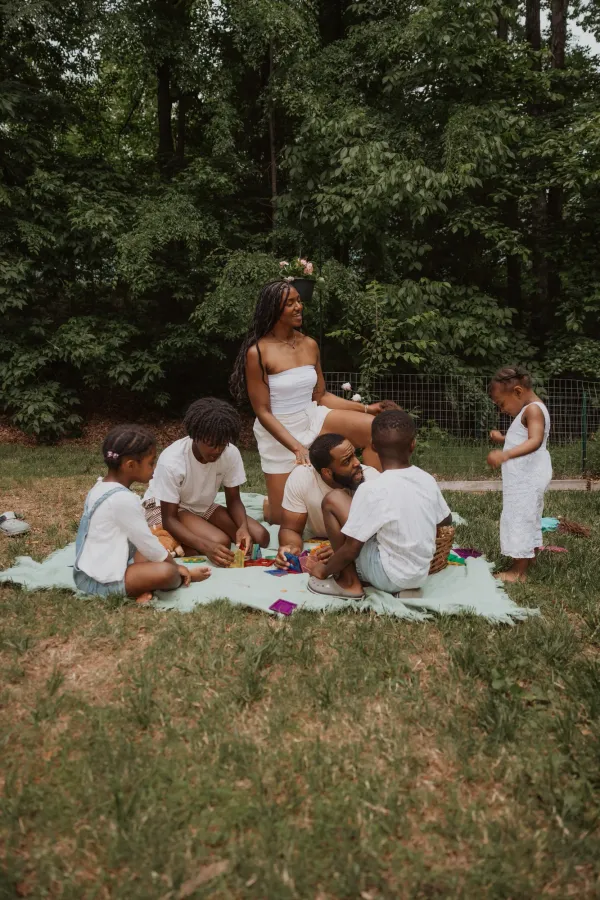 A family with children playing on a green blanket outdoors, surrounded by trees.